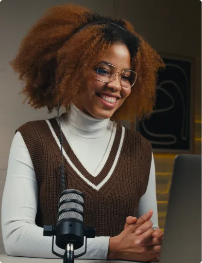 Smiling woman with curly hair and glasses wearing a brown vest and white turtleneck, sitting at a desk with a microphone and laptop.